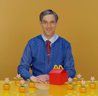 Alex Da Corte caracterizado como Mister Rogers junto a una caja de menú infantil y flores